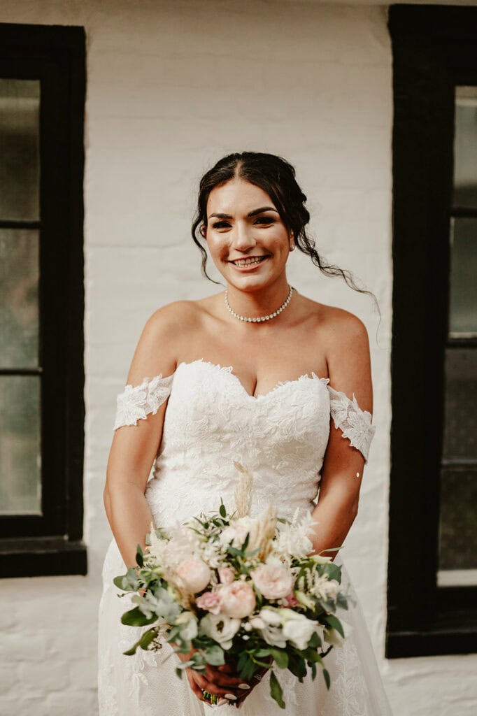 A bride with an off-the-shoulder lace wedding dress and a pearl necklace smiles while holding a bouquet of flowers, standing in front of a white brick wall with black-trimmed windows.