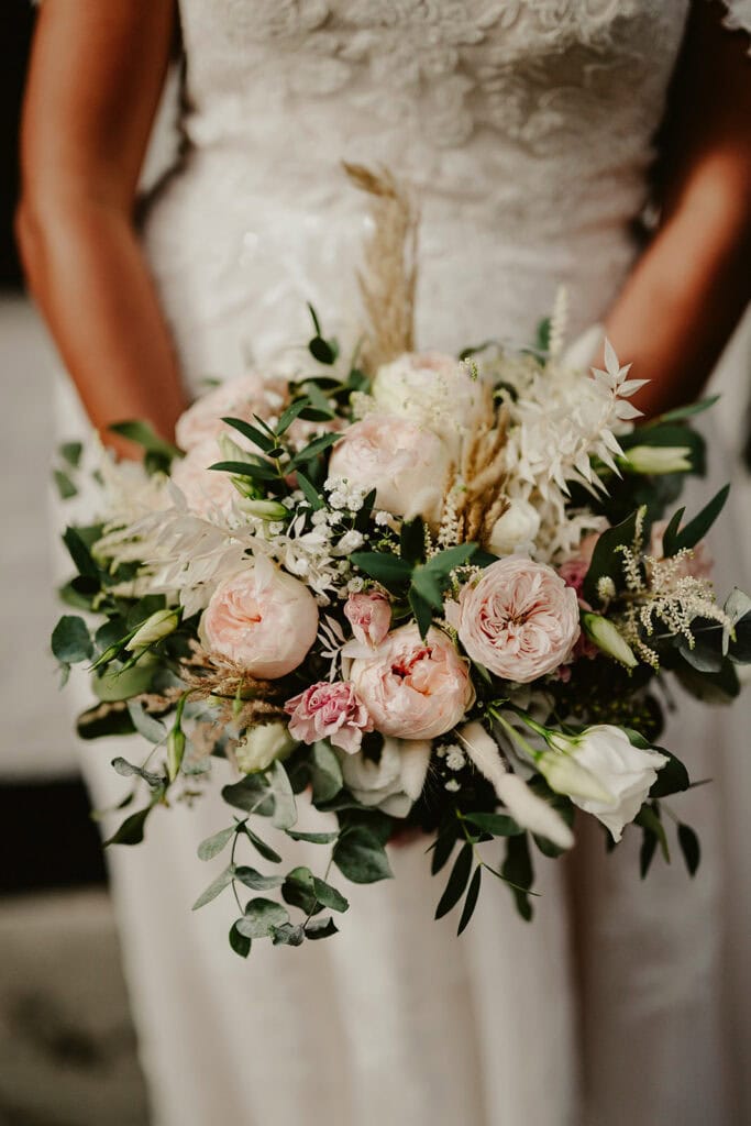 A bride in a white lace dress holds a bouquet of light pink and white flowers with green foliage.