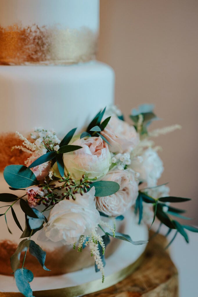 Close-up of a white tiered cake decorated with gold accents, adorned with pink and white flowers and green foliage.