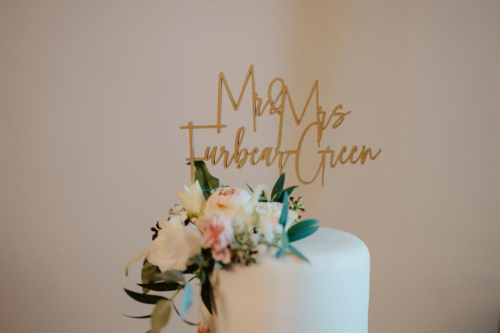 A white wedding cake topped with a gold "Mr & Mrs Everheart Green" sign and decorated with white and pink flowers.