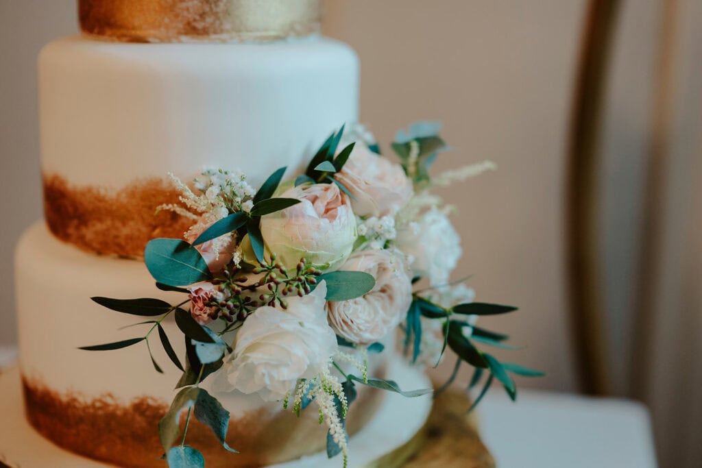 A three-tier cake with a gold accent is decorated with pink and white flowers and green leaves.