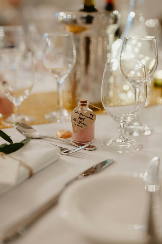 A formal table setting with a handwritten bottle of hot chocolate, various empty wine glasses, cutlery, and a folded napkin on a white tablecloth.