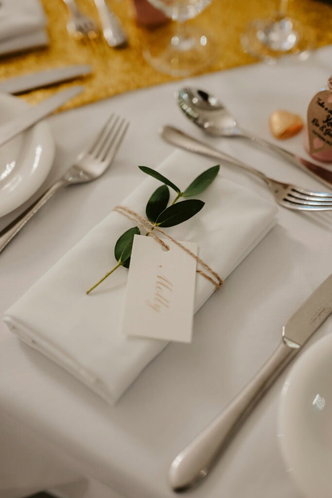 A neatly folded white napkin with a small green branch is tied with twine and placed on a table setting, featuring a name tag that reads "Maddy." Cutlery and a gold table runner are also visible.