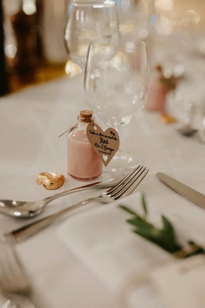 A close-up of a table setting with a fork, knife, wine glass, a small bottle with a heart tag, and wrapped chocolates.