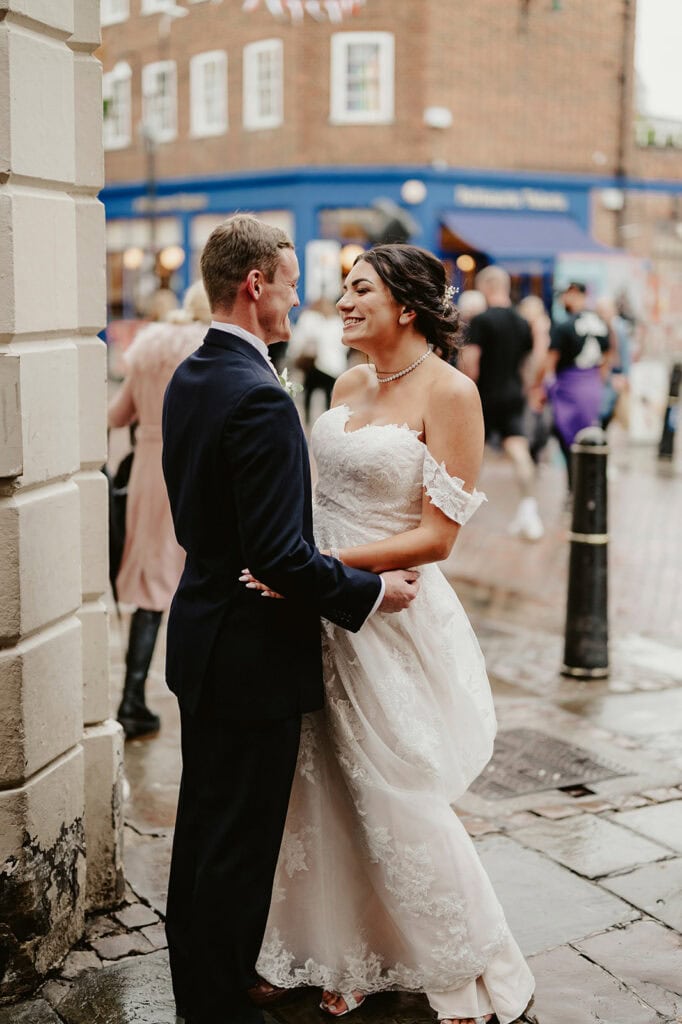 A bride and groom stand close together outside on a rainy day. The bride is in a white lace dress, and the groom is in a dark suit. People and buildings are visible in the background.