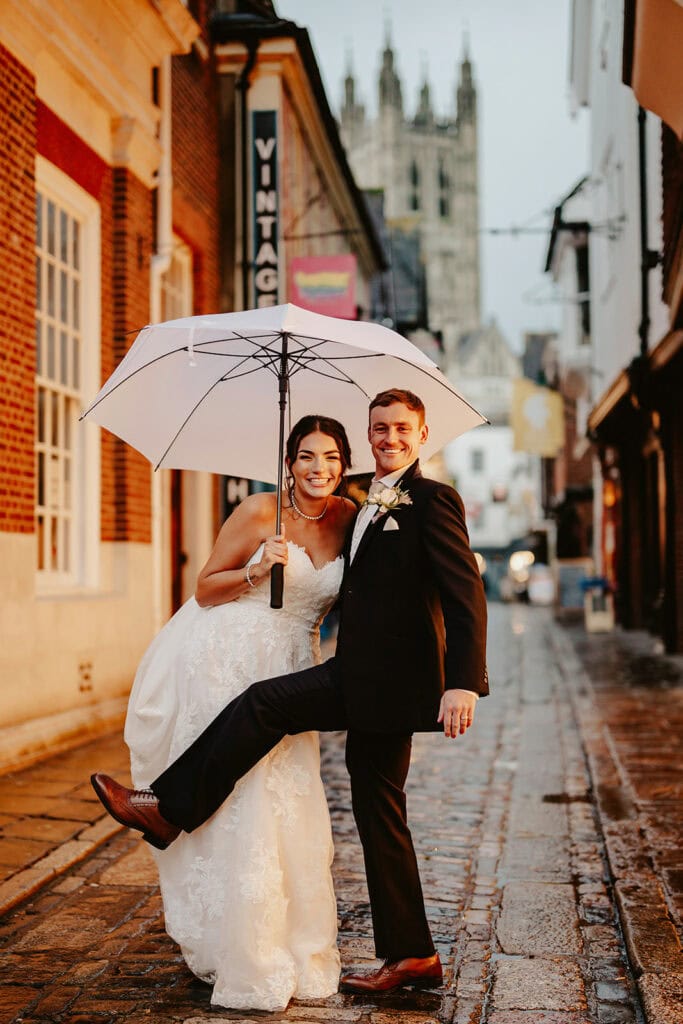 A bride and groom stand under a white umbrella on a wet cobblestone street. The groom lifts one leg playfully, while the bride smiles, with a tall historic building in the background.