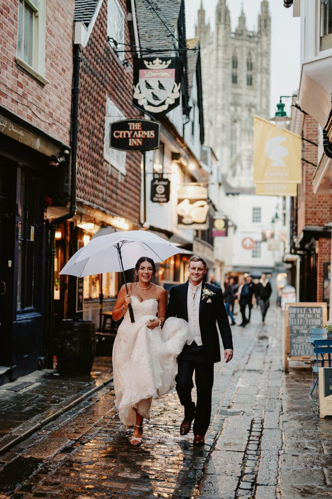 A bride and groom walk down a wet cobblestone street under an umbrella. Historic buildings and a cathedral tower are visible in the background. The bride holds her dress and the groom wears a suit with a boutonniere.