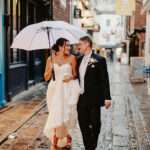 A bride and groom walk arm in arm on a wet cobblestone street, sharing a white umbrella. The bride holds her dress and wears white heels, while both smile, looking at each other.