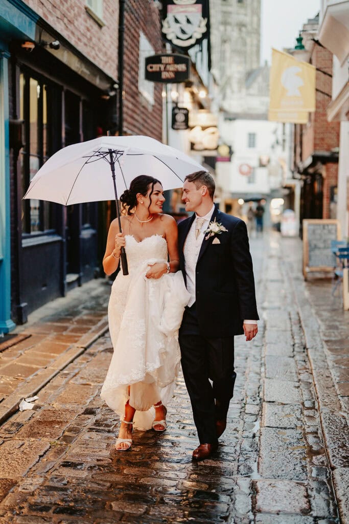 A bride and groom walk arm in arm on a wet cobblestone street, sharing a white umbrella. The bride holds her dress and wears white heels, while both smile, looking at each other.
