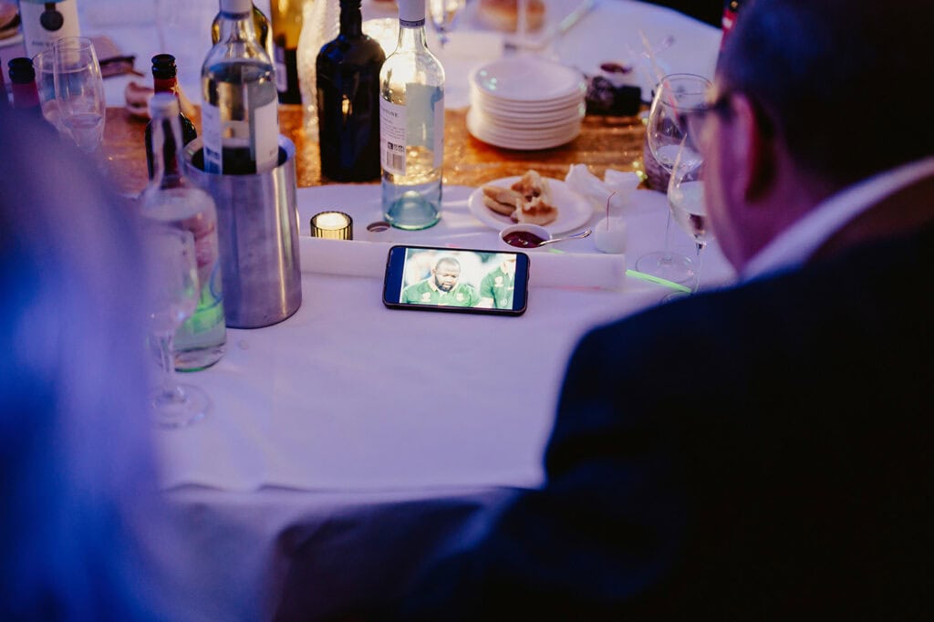 A person watches a video on a smartphone at a cluttered table with bottles, glasses, and plates.