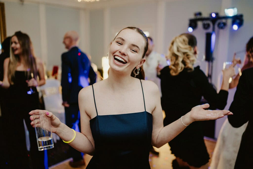 A smiling woman in a dark dress holds a drink and gestures with her other hand at a social gathering. Several people in the background appear to be mingling and talking.