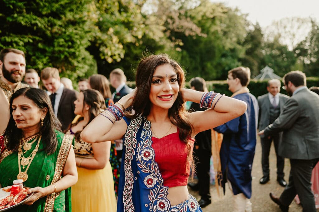 A woman in a colorful, traditional outfit smiles and poses with her hands near her head, surrounded by others in similar attire at an outdoor gathering.