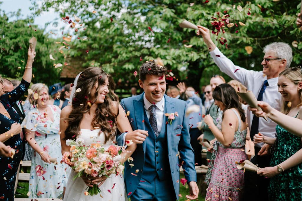 A smiling bride and groom walk down an aisle outdoors at bore place while guests throw flower petals in celebration. The bride holds a bouquet, and the groom wears a blue suit. Captured by a skilled wedding photographer in Kent, trees and greenery provide a stunning backdrop.