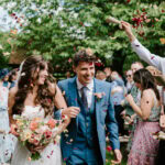 A smiling bride and groom walk down an aisle outdoors at bore place while guests throw flower petals in celebration. The bride holds a bouquet, and the groom wears a blue suit. Captured by a skilled wedding photographer in Kent, trees and greenery provide a stunning backdrop.