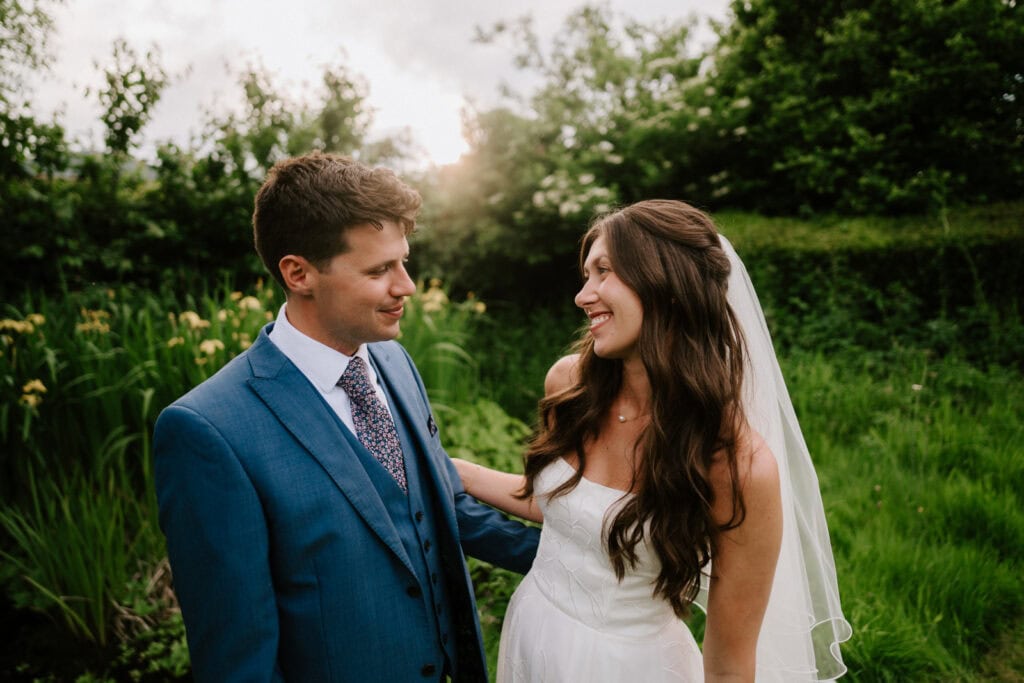 A bride and groom, captured perfectly by a Kent wedding photographer, smile at each other in a lush green garden. The groom dons a blue suit and tie, while the bride wears a white dress with a veil, surrounded by greenery and yellow flowers under the softly shining sun. Image by Pearce Wedding Photography.
