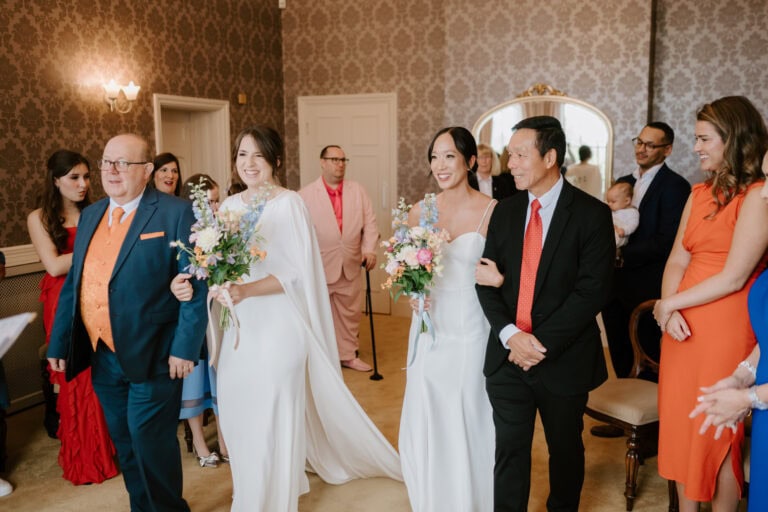 In an elegantly decorated room, two brides in white gowns make their way down the aisle of Aberdeen House, accompanied by individuals in suits. Holding beautiful floral bouquets, they are watched by guests in vibrant attire smiling warmly at this picturesque moment captured by a Thanet wedding photographer. Image by Pearce Wedding Photography.