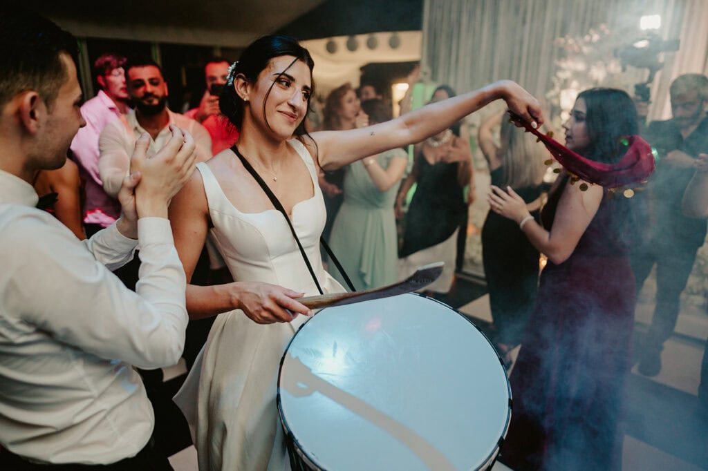 A woman in a sleeveless white dress enthusiastically plays a large drum as part of a lively dance celebration at Broome Park. She extends a red scarf to a smiling man in a white shirt beside her. People in the background join in the festive atmosphere, captured beautifully by the Canterbury wedding photographer amidst colorful lights and smoke effects. Image by Pearce Wedding Photography.