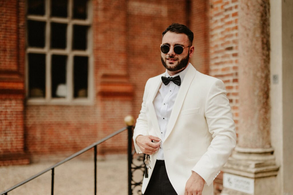 A man in a white suit jacket and bow tie stands confidently outdoors in front of a brick building. He is wearing dark sunglasses.