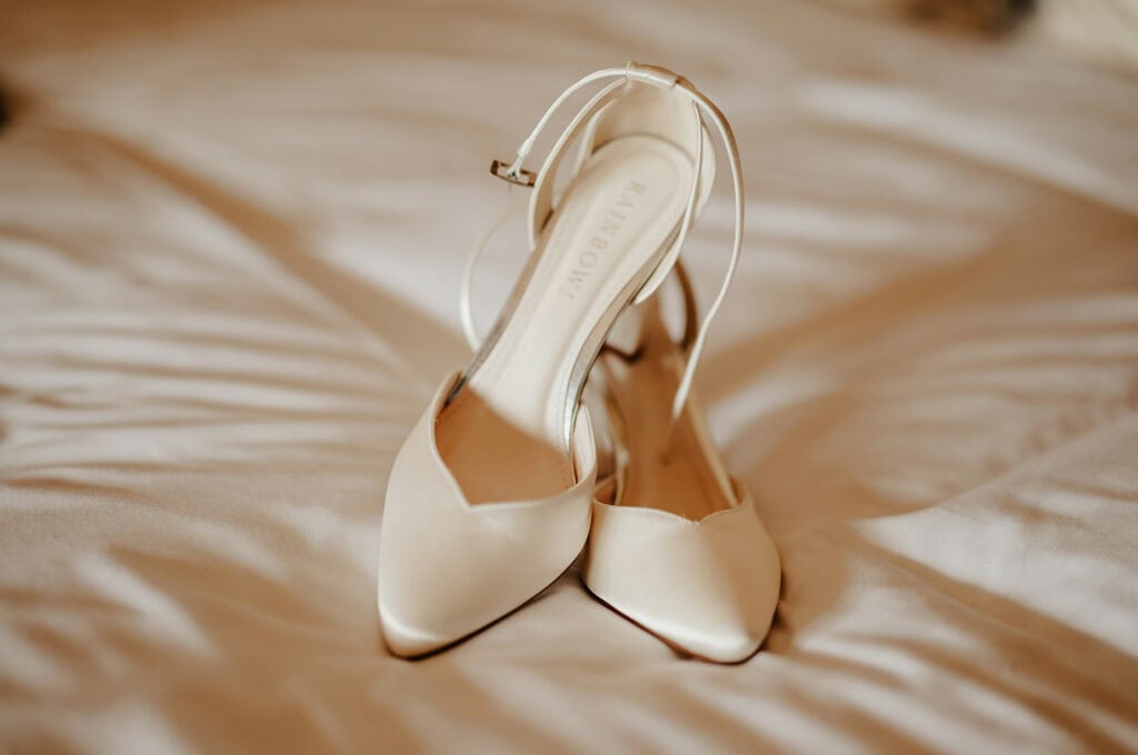 A pair of white pointed-toe high heels with ankle straps are placed on a white fabric surface.