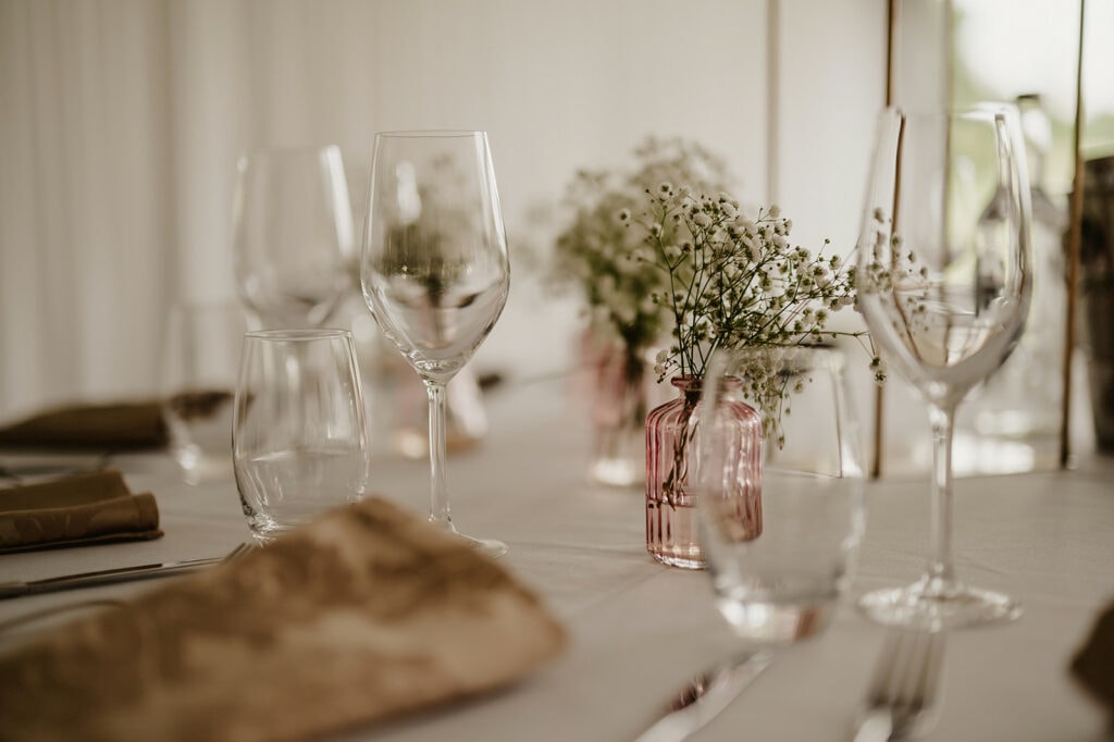 A close-up of a dining table set with wine glasses, water glasses, small pink vases with white flowers, and beige napkins, under soft lighting.