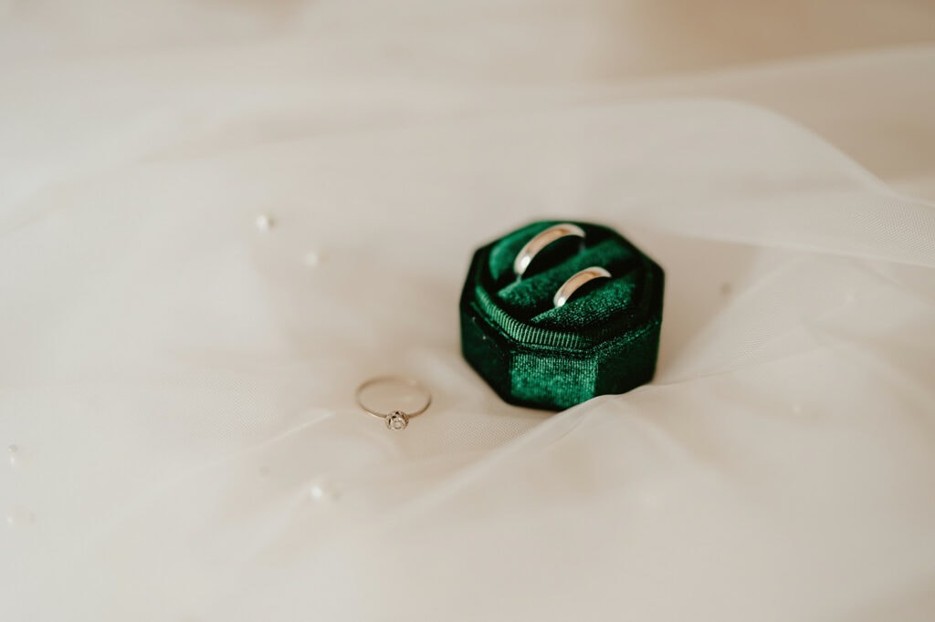 Two silver rings rest in a green velvet ring box next to a single diamond ring on sheer white fabric.
