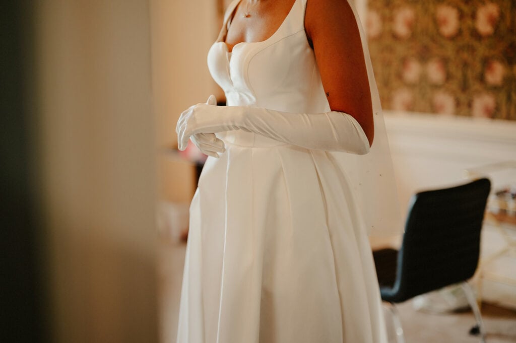 A woman in a white wedding dress and white gloves stands indoors near a chair.