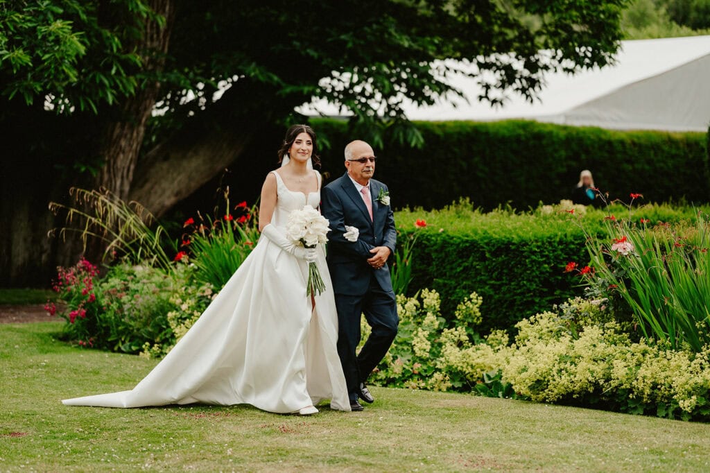 A bride in a white dress holding a bouquet walks arm-in-arm with an older man in a suit through a garden with lush greenery and colorful flowers.