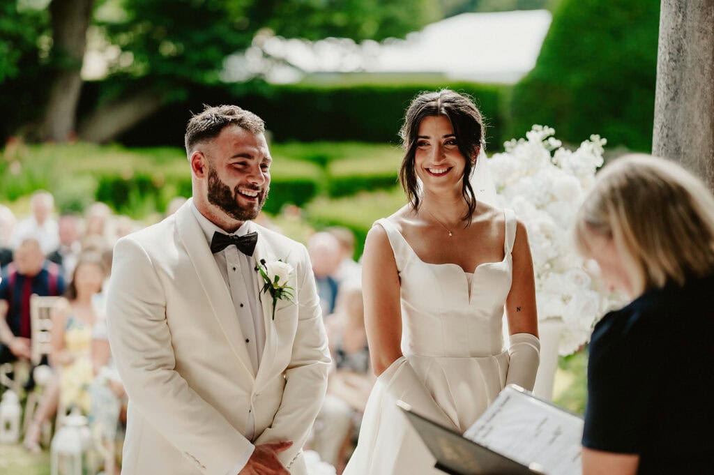 A couple in formal wedding attire stands smiling during their outdoor wedding ceremony at Broome Park in Kent, with a woman officiating the event. The background features greenery and seated guests.