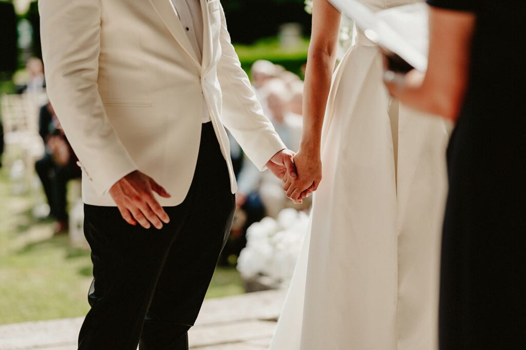 A couple dressed in formal attire holds hands during an outdoor wedding ceremony.