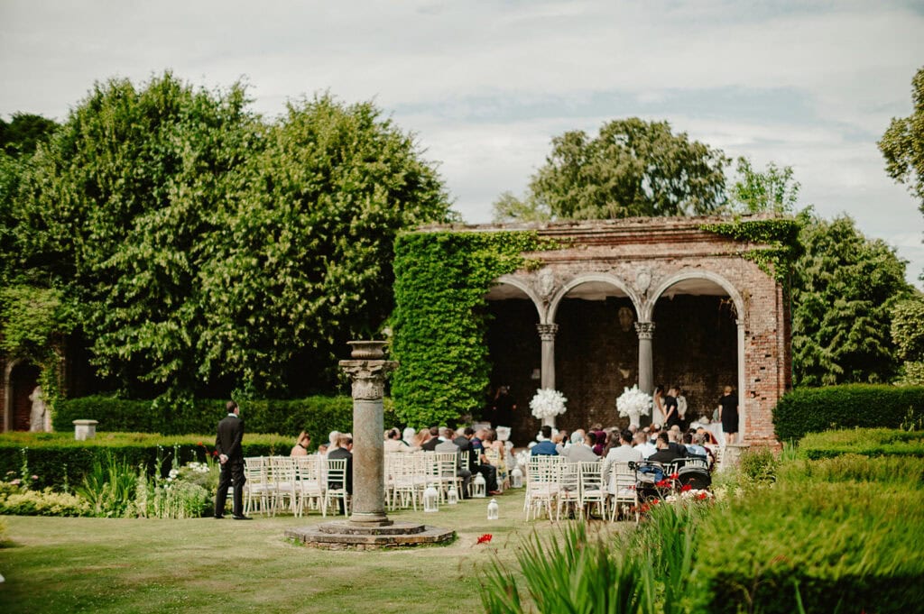 Outdoor wedding ceremony with guests seated on white chairs, facing a bride and groom under a columned, ivy-covered structure, surrounded by greenery and garden hedges.