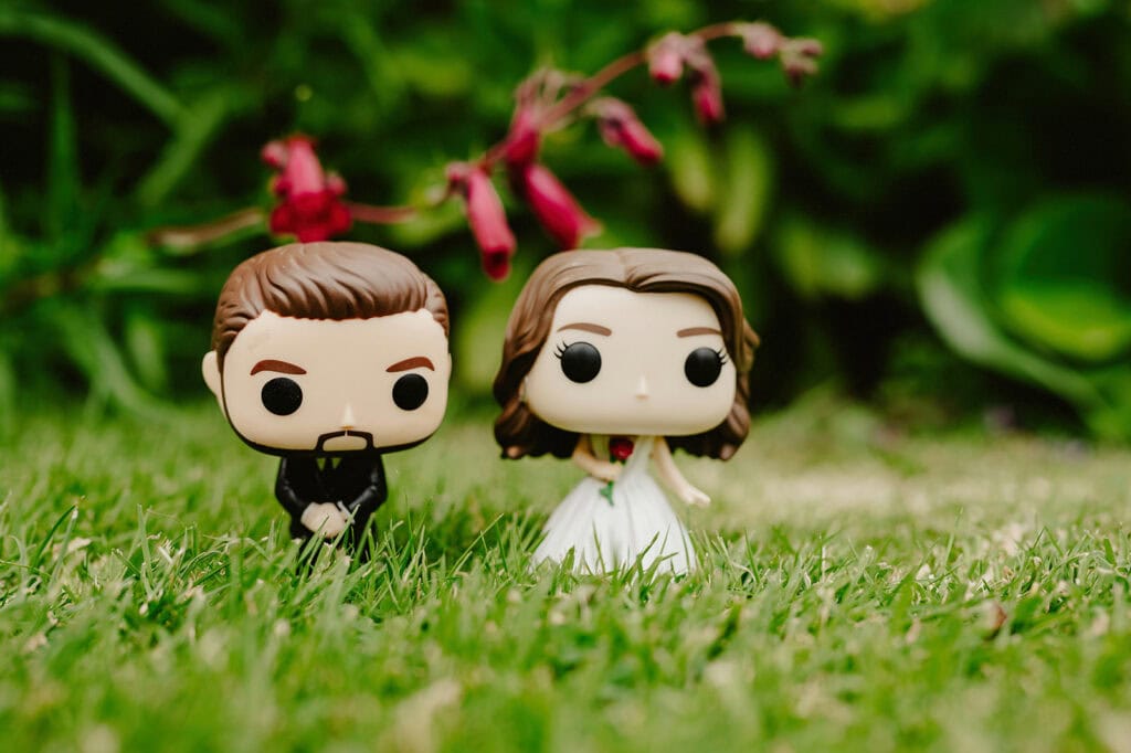 Two Funko Pop! vinyl figures of a bride and groom are positioned on vibrant green grass with blurred flowers and foliage in the background.