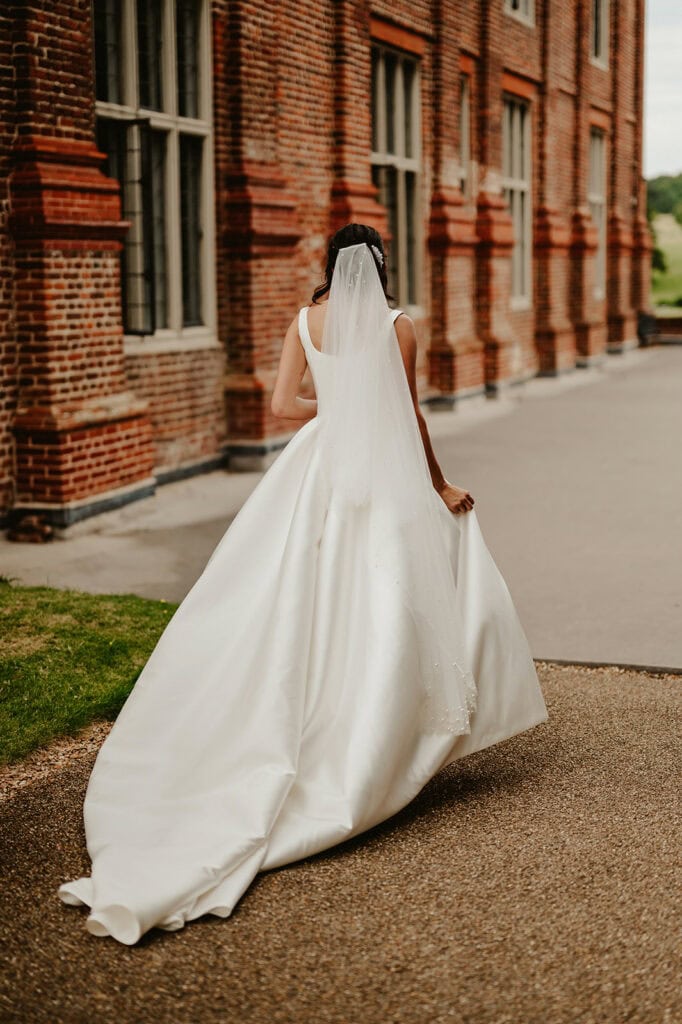A bride in a white wedding dress and veil walks alone along a pathway next to a brick building.