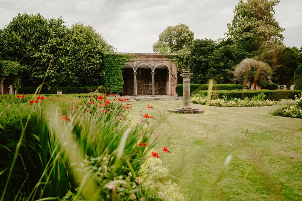 A garden at Broome Park, featuring a stone structure of arches covered in ivy, surrounded by lush green bushes, red flowers, and manicured lawns under a cloudy sky—an enchanting setting perfect for a wedding in Kent.