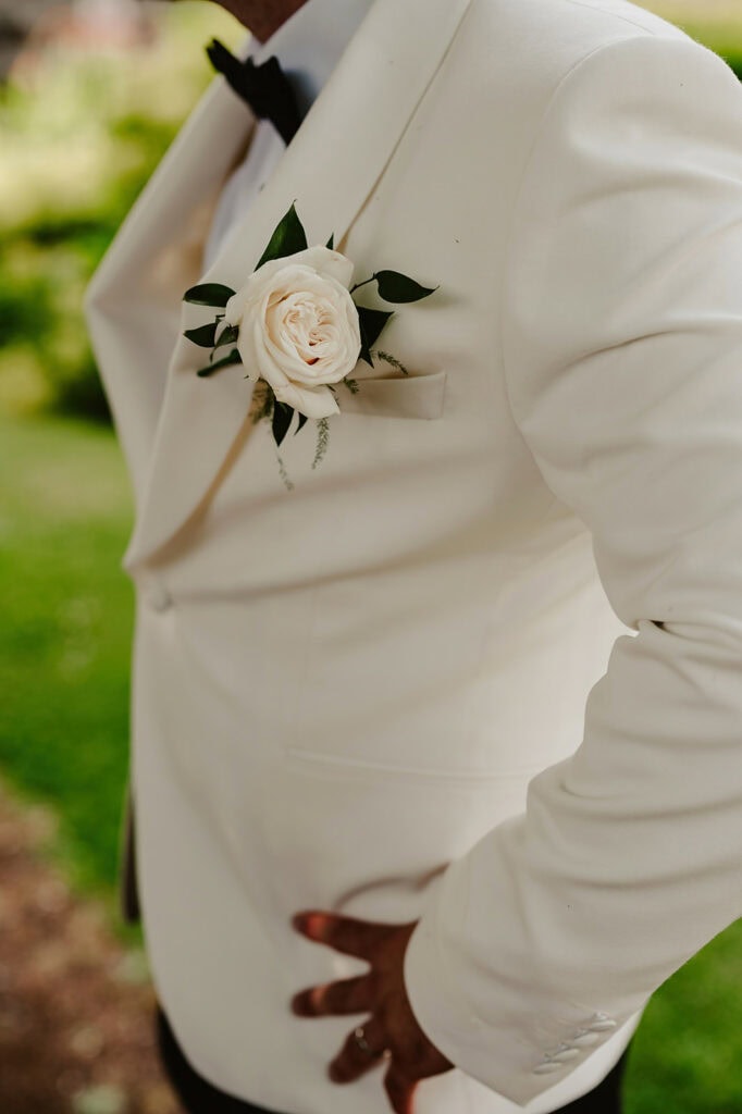 Person in a white tuxedo jacket with a white rose boutonniere pinned to their lapel, standing outdoors.