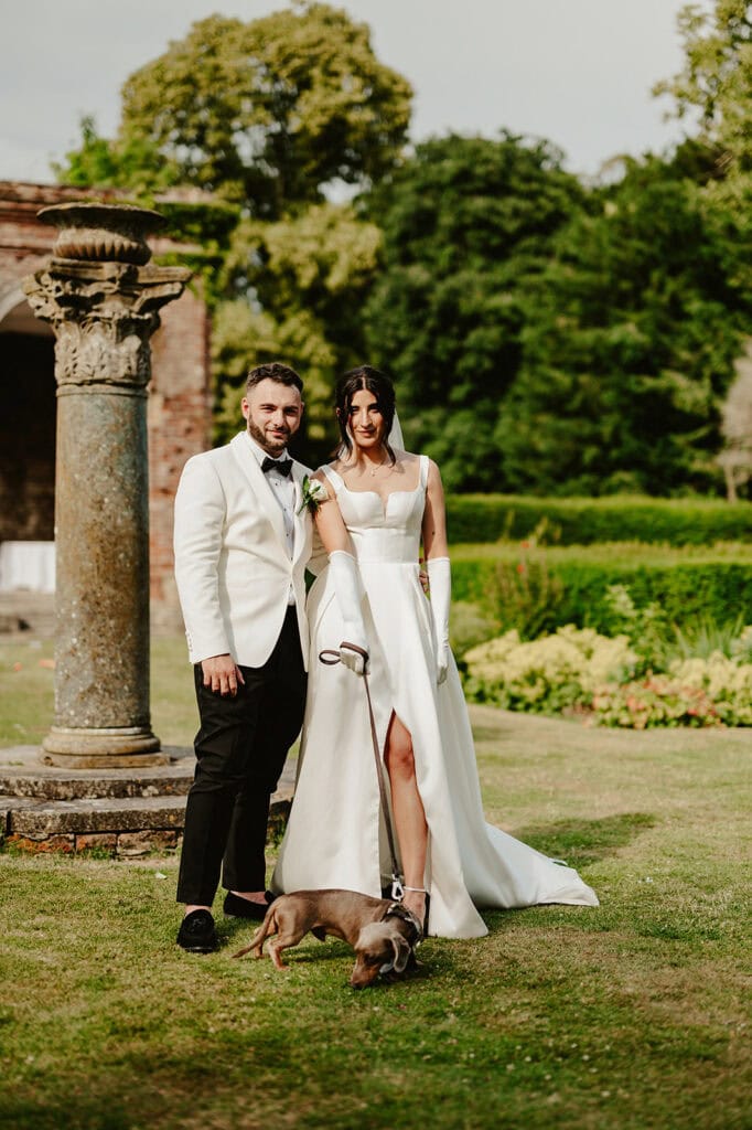 A bride and groom stand outdoors on a grassy area at Broome Park, with the groom wearing a white jacket and black pants, and the bride in a white dress with a slit. They are both smiling and holding a leash attached to a small brown dog at their feet. Trees and shrubs are in the background, captured beautifully by their Canterbury wedding photographer. Image by Pearce Wedding Photography.