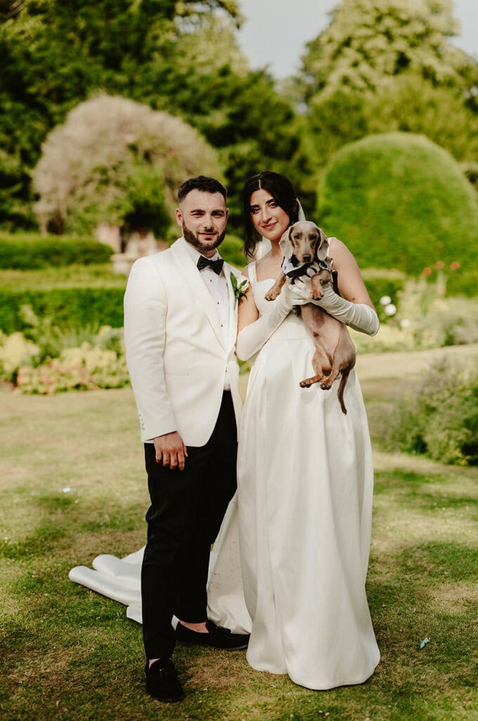 A bride and groom in formal attire stand outdoors at Broome Park in Kent, with the bride holding a small dog.