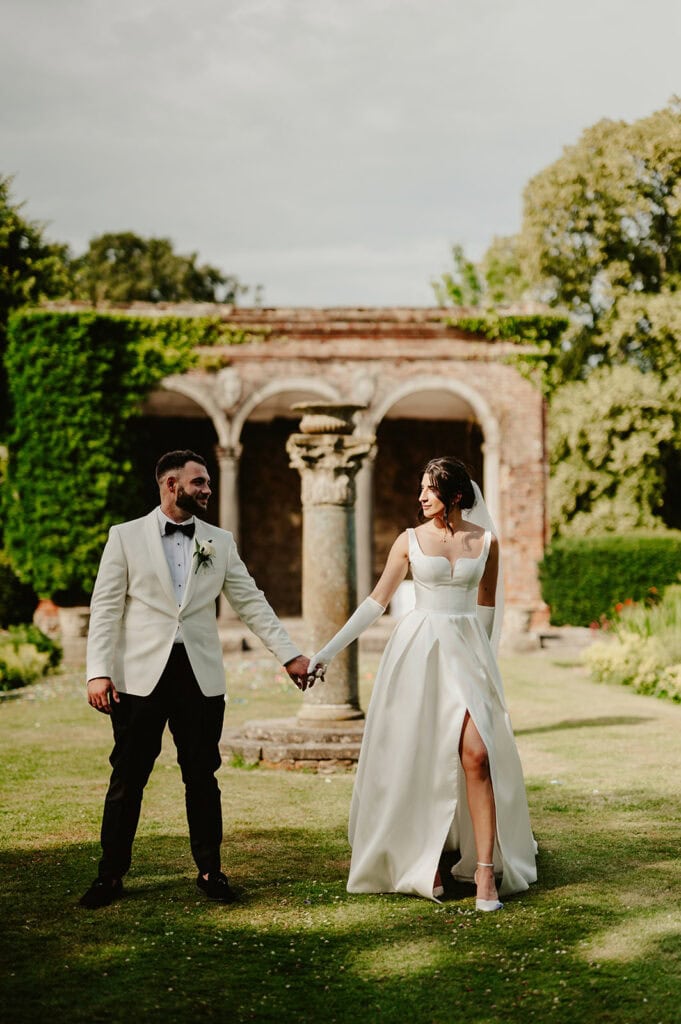 A bride and groom hold hands while walking on a grassy lawn. The bride is in a white dress and the groom is in a white jacket and black pants. An ancient stone structure is in the background.
