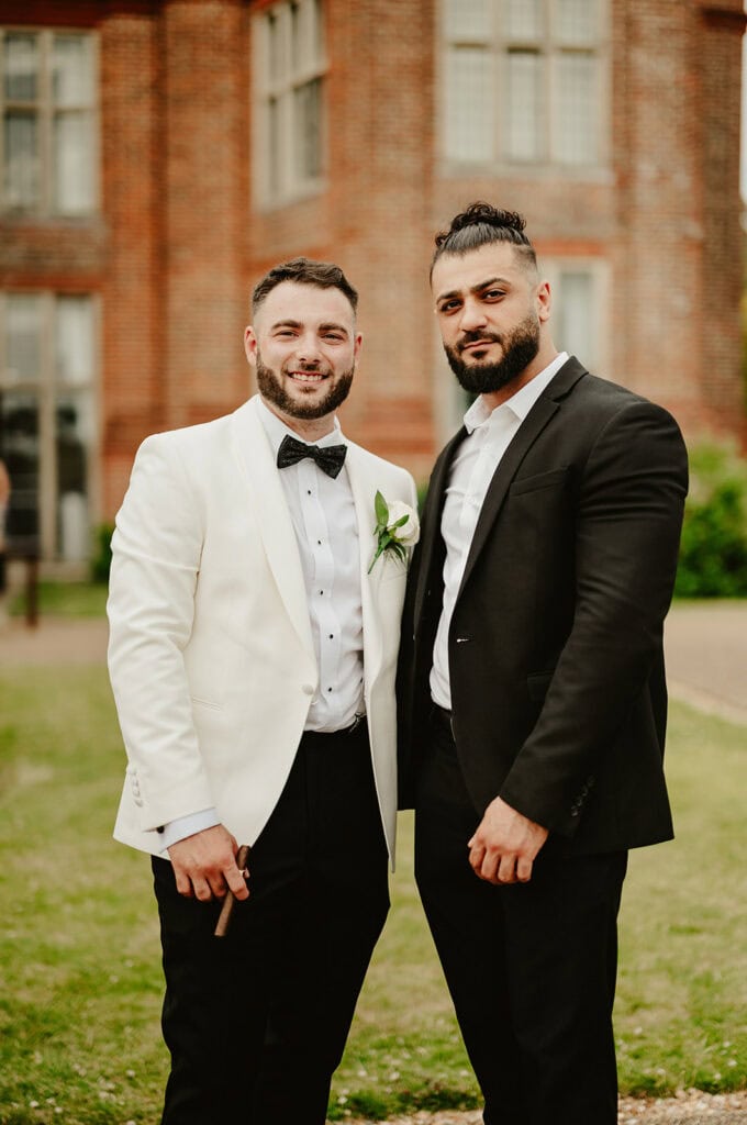 Two men in formal attire stand side by side outdoors. One wears a white tuxedo jacket with a black bow tie and the other wears a black suit. A brick building is in the background.