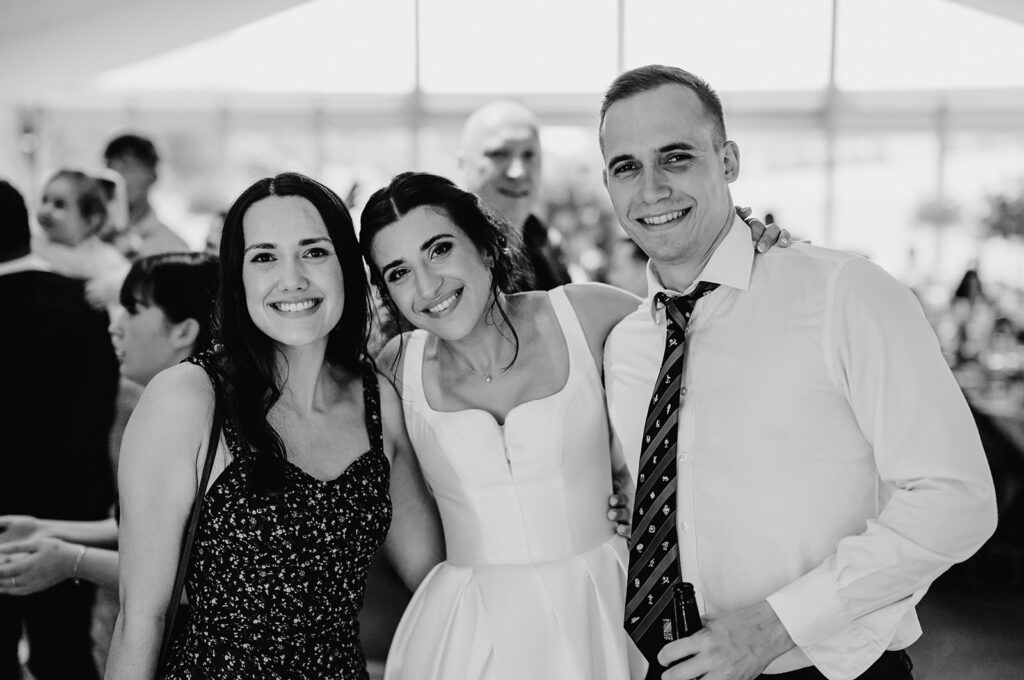 Three people stand closely together, smiling for a photo. The woman in the center is wearing a white dress, likely a wedding gown, while the other two wear formal attire. Blurred people are visible in the background.