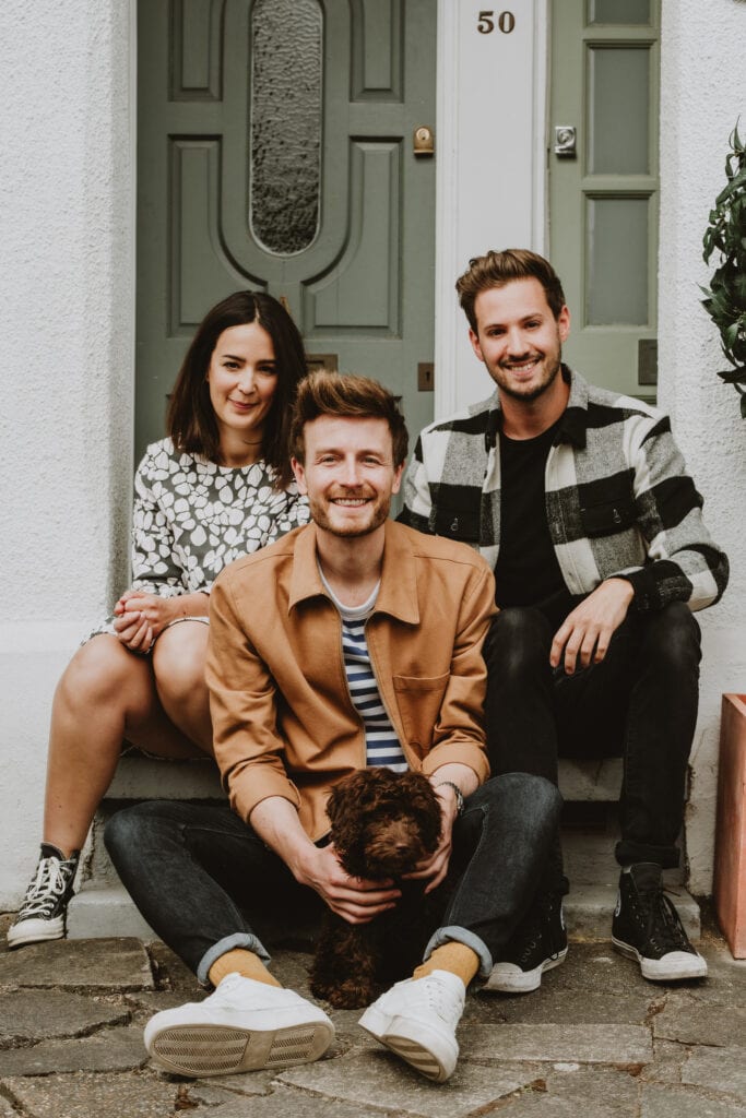 Three people sitting on the steps of a house with a green door numbered 50. One person is holding a small brown dog. They are all smiling and appear relaxed.