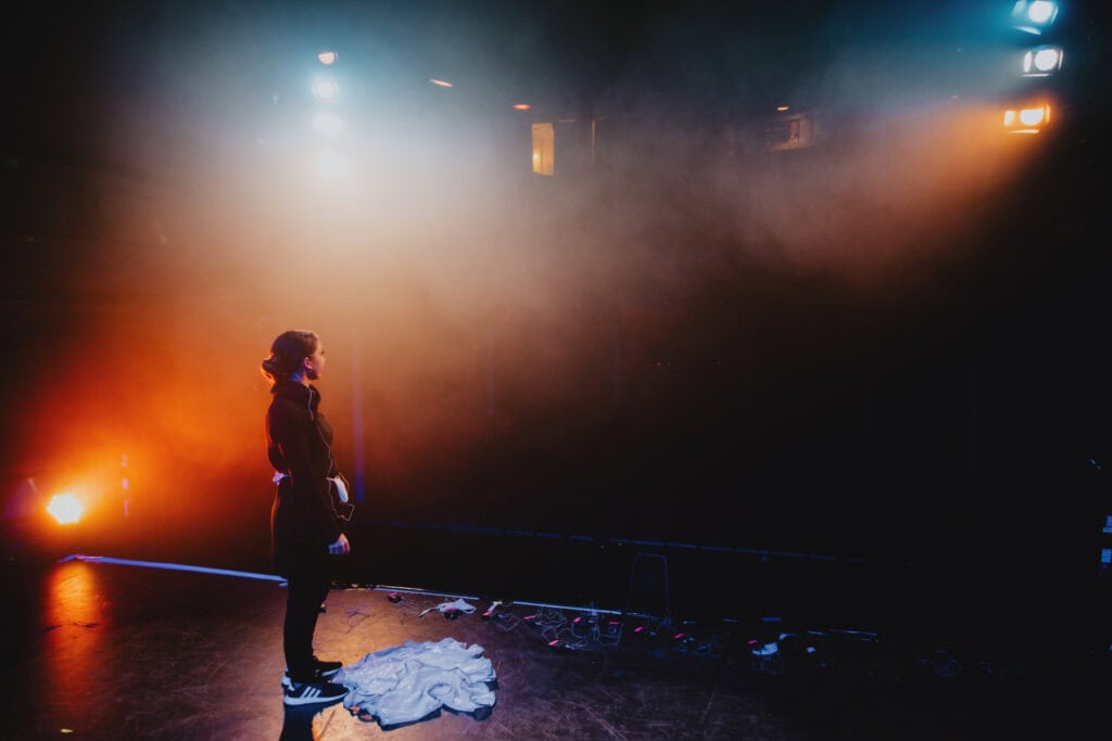 Person standing on a dimly lit stage with bright spotlights overhead, facing the audience, wearing casual clothes. There are scattered items, including cables and fabric, on the floor behind them. A renowned music photographer from London captures this intimate moment amidst the chaos of a Kent venue.