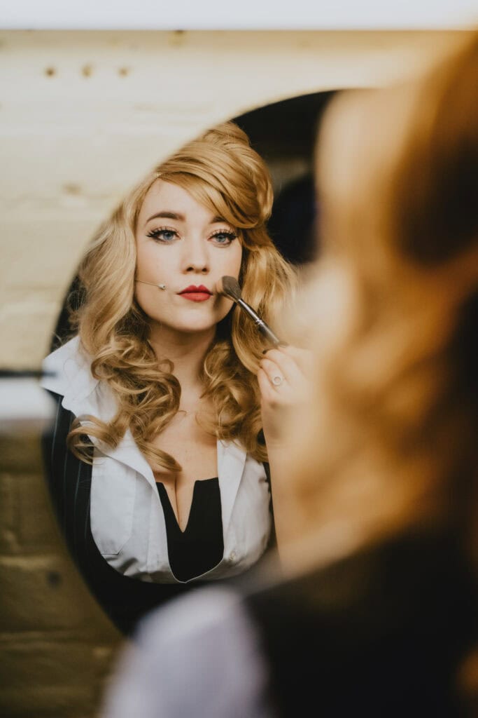 A woman with curly blonde hair applies makeup, looking into a mirror. She is holding a brush near her face and wears a white blouse with a black corset.