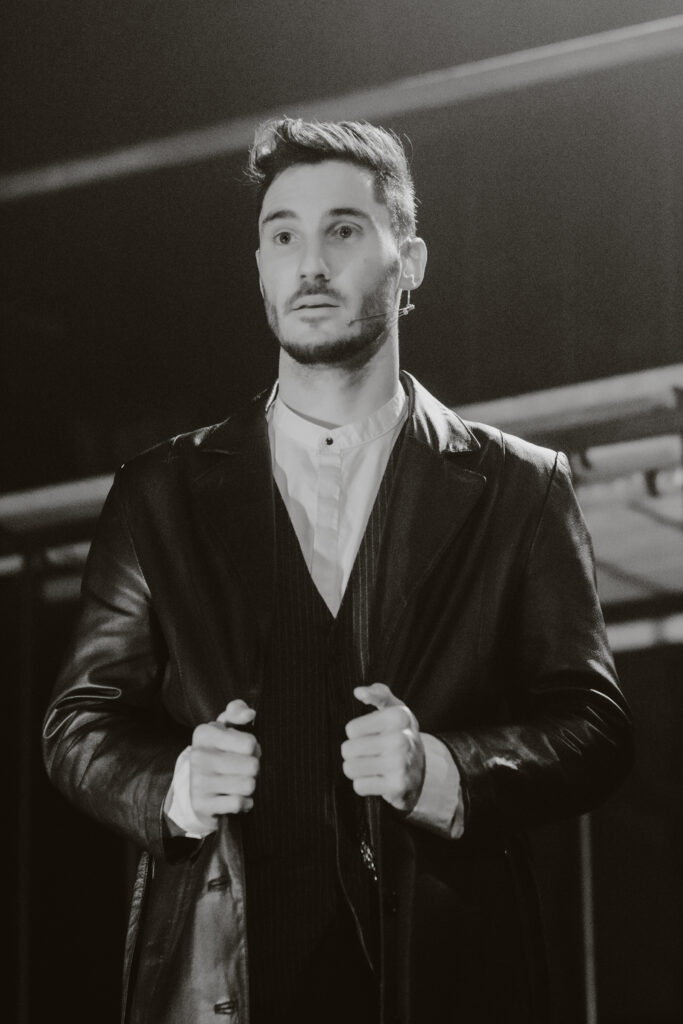 A man in a leather jacket and white shirt stands with his hands on his jacket lapels, exuding the cool demeanor of a Kent-based music photographer. He appears to be looking off into the distance. The image is in black and white.