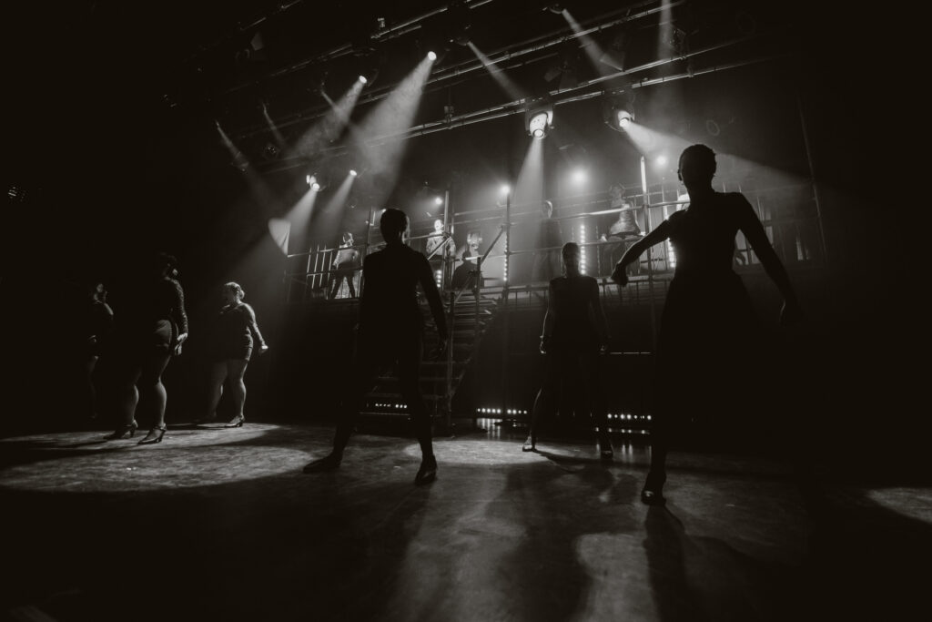 A group of performers on stage with dramatic lighting, some standing and others mid-movement. A staircase and elevated platform with additional people are visible in the background, capturing the essence of a dynamic moment as seen through the lens of a London music photographer.