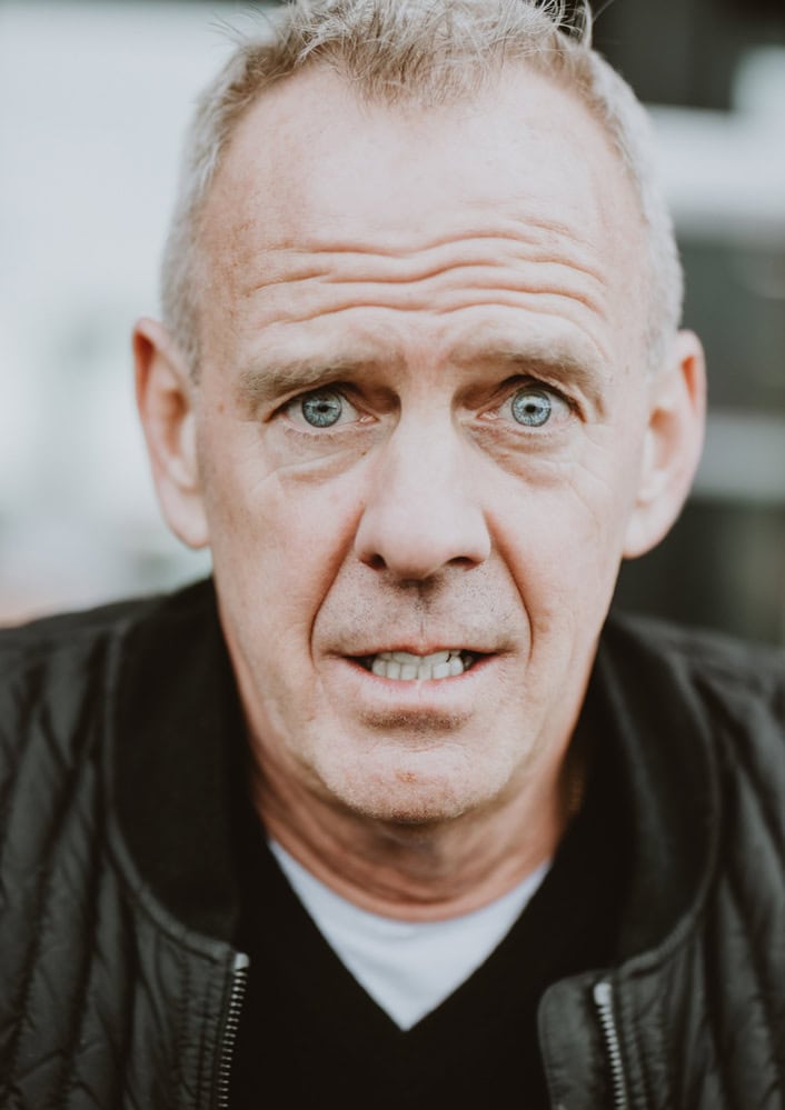 A middle-aged man with short hair and blue eyes looks at the camera with a slight frown. He is wearing a black jacket and a black shirt, giving him the appearance of a seasoned music photographer. The background is blurred, adding an air of mystery to this portrait taken in London.