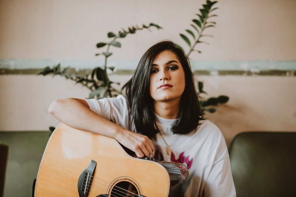 A music photographer captures a woman with shoulder-length dark hair holding an acoustic guitar. She wears a white shirt and sits in front of a plant, looking directly at the camera. This serene moment was photographed in London, blending urban charm with musical passion.