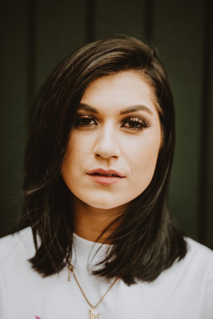 A woman with straight, shoulder-length dark hair, wearing a white top and a necklace, looking directly at the camera with a neutral expression. Background is dark and plain.