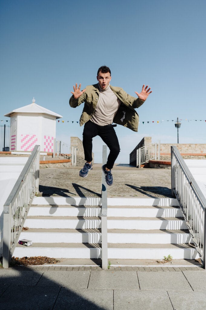 A person in a green jacket and black pants is jumping over a set of stairs outdoors on a sunny day, with arms raised for balance. A white building and colorful flags are visible in the background.