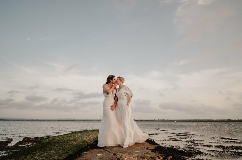 Two people in wedding dresses stand on a rocky shore by the water, sharing a gentle kiss under a cloudy sky, with the ferry house visible in the distance.