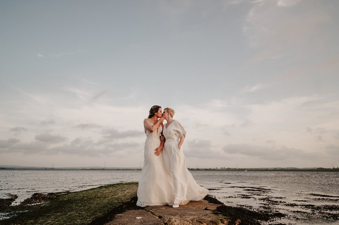 Two women in wedding dresses stand on a stone path by the water, sharing an intimate moment. The cloudy sky above adds to the serene and romantic atmosphere, beautifully captured by a Kent wedding photographer celebrating their same-sex union. Image by Pearce Wedding Photography.
